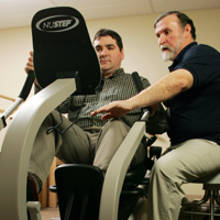 Picture of a male Physical Therapist helping assist a male patient on an excessive machine.